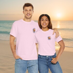 May include: Two people wearing light pink t-shirts with a small circular logo on the left chest. The logo features a beach scene with the words "Hooked & Salty". Both are wearing blue jeans. The background is a beach with an ocean and a sunset.