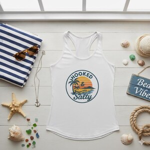 May include: White tank top with a circular graphic that reads "Hooked & Salty" with a beach scene. The image includes a striped towel, sunglasses, starfish, seashells, and a small wooden sign that says "Beach Vibes".