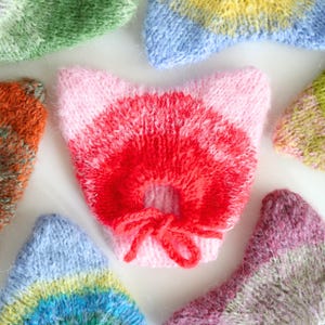Könnte beinhalten: Nahaufnahme von mehreren handgefertigten, gestrickten katzenförmigen Artikeln in verschiedenen Farben. Das zentrale Stück ist rosa und rot mit einer kleinen Schleife. Andere Teile sind blau, grün und lila, mit gestreiften und einfarbigen Mustern. Dies sind wahrscheinlich Katzenspielzeuge oder -accessoires.
