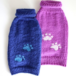 Puede incluir: Dos suéteres tejidos para mascotas, uno en azul profundo con patrones de huellas en azul claro y otro en morado vibrante con patrones de huellas en blanco. Ambos suéteres cuentan con cuello de tortuga acanalado y dobladillo acanalado.