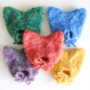 May include: Five fuzzy, hand-knitted bonnets in distinct colors: a variegated green, a bright blue, a mottled red, a purple and blue blend, and a solid yellow. Each bonnet features a pointed top and a small tie closure at the chin area.
