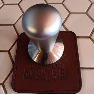 May include: A silver coffee tamper with a round handle rests on a brown leather coaster. The coaster has the text "THE ROAST OFFICE" and "HOT COFFEE & COLD RESULTS" embossed on it. The background is a white hexagonal tile.