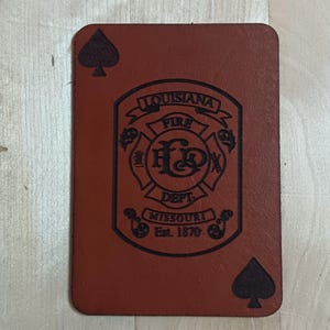May include: A brown leather patch with a black spade on each corner. The center of the patch features a black and white emblem with the text "Louisiana Fire Dept. Missouri Est. 1870."