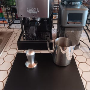 May include: A stainless steel Gaggia Classic espresso machine and a coffee grinder sit on a countertop. A silver tamper, a stainless steel milk frothing pitcher, and a black mat are in the foreground. The machine has a black portafilter.