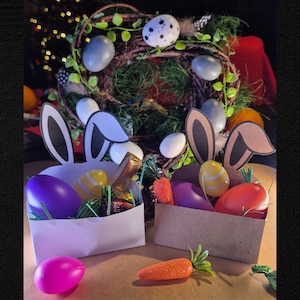 May include: Two Easter baskets with bunny ears, filled with colorful eggs and candy. One basket is white, the other is brown. A decorative wreath with eggs and greenery is in the background. Orange carrots are placed in front of the baskets.