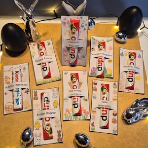 May include: An assortment of Easter-themed gifts featuring chocolate bars with hazelnut filling. Each bar is presented on a decorative card with various Easter greetings and illustrations, including bunnies and eggs. The color scheme includes red, white, and brown.