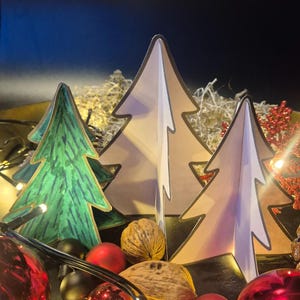 Puede incluir: Una escena festiva navideña con tres árboles de Navidad de papel en tonos verdes y beige. Los árboles están decorados con luces de cadena, adornos rojos y negros y nueces, creando un ambiente cálido y acogedor.