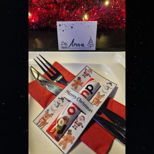 Könnte beinhalten: Eine festliche Weihnachtsdekoration mit einer roten Serviette, Besteck und einer Platzkarte mit dem Namen "Anna". Ein Schokoladen-Leckerbissen liegt auf einer dekorativen Karte mit "Merry Christmas" und festlichen Illustrationen.