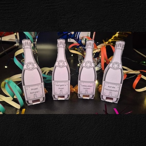 May include: Four champagne bottle-shaped paper cutouts with text in German. Each bottle has a star and a label with phrases like "Prost!" and "Auf uns!" in black ink. Colorful streamers and a glass are in the background.