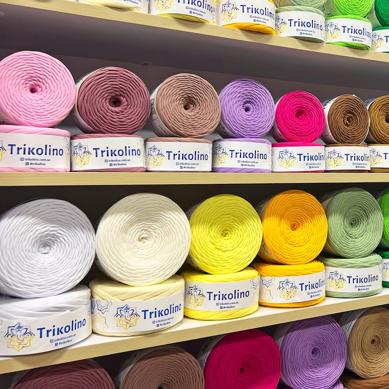May include: Shelves filled with colorful yarn balls in various shades, including pink, purple, yellow, and green. Each ball is labeled with the brand name "Trikolino". The yarn is neatly arranged, showcasing a variety of colors for crafting projects.
