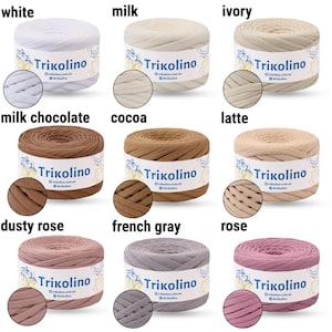 May include: A collection of Trikolino yarn in various colors, including white, milk, ivory, milk chocolate, cocoa, latte, dusty rose, French gray, and rose. Each skein is labeled with the Trikolino logo and color name. The yarn is displayed in a neat arrangement.