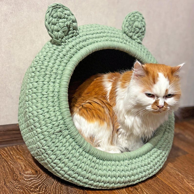 May include: A handmade, light green cat bed with ear-shaped details. The round bed is made of thick, woven material. An orange and white cat is inside the bed, looking towards the viewer.