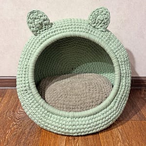 May include: A mint green, crocheted cat bed with a round shape and two ear-shaped accents on top. The bed has a circular opening and a grey, plush cushion inside. The bed is placed on a wooden floor.