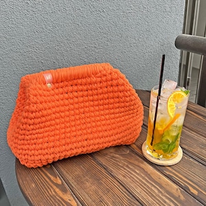 Può includere: Una pochette all'uncinetto arancione è appoggiata su un tavolo di legno accanto a un bicchiere alto di una bevanda rinfrescante con ghiaccio, una fetta di limone e una cannuccia. La borsa è realizzata con filato spesso e strutturato.
