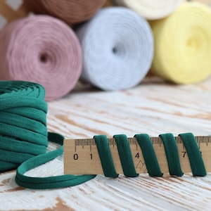 May include: A close-up of various spools of yarn in pastel colors, including pink, white, and yellow. A wooden ruler displays a dark green fabric ribbon, with measurements in inches. The yarn is likely for crafting projects.
