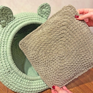 May include: A light green, handmade crochet pet bed with ear-shaped accents. A removable, textured, light brown pillow is held nearby. The bed is round with a large opening. The pillow is square.