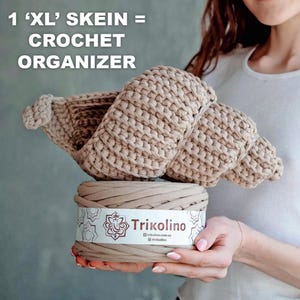 May include: A beige crochet organizer sits atop a skein of beige yarn. The text in the image reads: "1 'XL' SKEIN = CROCHET ORGANIZER". The yarn is labeled "Trikolino". The crochet organizer is made of thick, woven yarn.