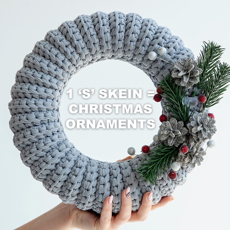 May include: A gray, knitted Christmas wreath with decorative pinecones, red berries, and evergreen sprigs. The wreath is held by a person. Text on the wreath reads: "1 'S' SKEIN = CHRISTMAS ORNAMENTS."