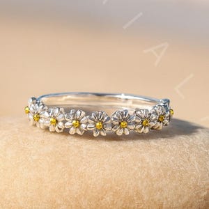 May include: A silver ring with a daisy flower design. The ring features small white petals and yellow centers, set in a band. The ring is displayed on a smooth, light brown stone, with a sandy beach background.
