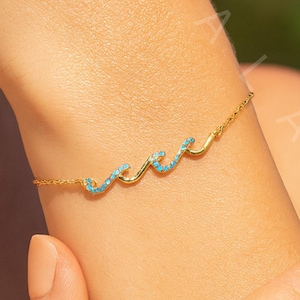 Pode incluir: Uma pulseira dourada com um design de onda e pedras de cor turquesa. A pulseira é usada no pulso, mostrando sua delicada corrente e o padrão de onda curvada. As pedras turquesas adicionam um toque de cor.