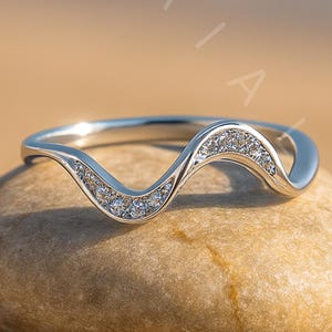 May include: A silver-colored ring with a wave design, set with small, clear gemstones. The ring sits on a smooth, light brown stone, with a blurred beach and ocean background.