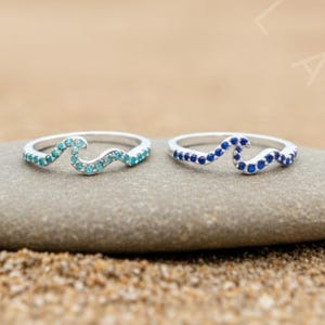 Può includere: Due anelli d'argento a forma di onda, uno con pietre turchesi e verde acqua, l'altro con pietre blu, esposti su una pietra grigia. Gli anelli sono ambientati su una spiaggia sabbiosa e un oceano.