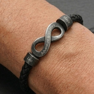 Op de afbeelding: Zwarte gevlochten leren armband met een zilverkleurig oneindigheidssymbool en twee zilveren kralen. Het oneindigheidssymbool is gegraveerd met de woorden "Adventure & Journey". De kralen zijn gegraveerd met de namen "Leo" en "Maya".