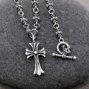 Pode incluir: Um colar prateado com um pendente de cruz e uma corrente decorativa. A cruz tem um design detalhado com bordas curvas e um elemento circular central. O colar é exibido em uma pedra cinza, com uma praia e o mar ao fundo.