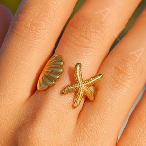 Puede incluir: Un anillo dorado con un diseño de concha y estrella de mar. La concha está a un lado del dedo y la estrella de mar al otro. El anillo se lleva en un dedo con esmalte de uñas rosa claro.