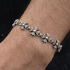 Op de afbeelding: Zilverkleurige armband met een herhalend patroon van gestileerde kruisachtige bloemmotieven. De armband wordt om de pols gedragen, waarbij de ingewikkelde details van het metaalwerk worden benadrukt. Een klassieke, tijdloze stijl.