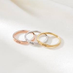 Può includere: Tre anelli personalizzati in oro rosa, argento e oro, ognuno inciso con il nome "Ella". Gli anelli sono presentati su una superficie bianca e setosa, che mette in risalto il loro design delicato e le diverse finiture metalliche.