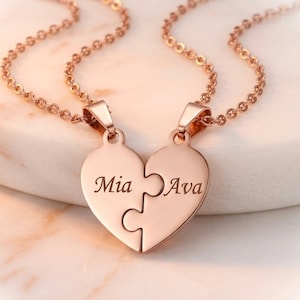 Peut inclure: Collier en or rose avec deux pendentifs en forme de cœur qui s'emboîtent. Chaque pendentif porte le nom "Mia" et "Ava", et est suspendu à une fine chaîne. Le collier est présenté sur une surface claire.