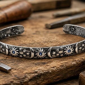May include: A silver cuff bracelet with celestial designs, including suns, moons, and stars, and small blue gemstones. The bracelet is set against a wooden surface with tools in the background, suggesting a handcrafted item.