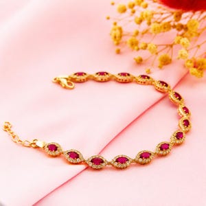 May include: Gold-toned bracelet featuring oval-shaped red gemstones surrounded by small, clear stones. The bracelet is displayed on a pink fabric, with a delicate clasp and adjustable chain. The jewelry is a classic design.