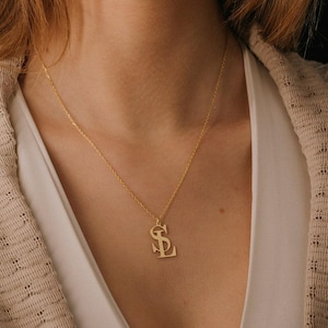 May include: A gold-colored pendant necklace featuring the intertwined letters "SL". The pendant hangs from a delicate chain, resting against a person's chest. The necklace is a simple yet elegant piece of jewelry.