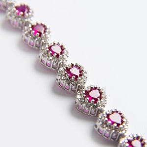 Puede incluir: Primer plano de una pulsera plateada con una serie de piedras preciosas redondas de color rubí. Cada piedra preciosa está rodeada de piedras más pequeñas y transparentes, creando un efecto de halo. La pulsera está sobre un fondo blanco.