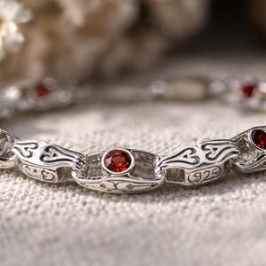 May include: A silver bracelet with a series of ornate links, each set with a round, red gemstone. The bracelet is made of silver and features a decorative pattern. The clasp is visible, and the bracelet is displayed on a textured surface.