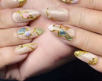 May include: A set of almond-shaped, glittery beige nails with gold accents. Each nail features a unique design with gold lines and colorful, mosaic-like patterns in shades of pink, blue, green, and gold. Some nails have small gold beads.