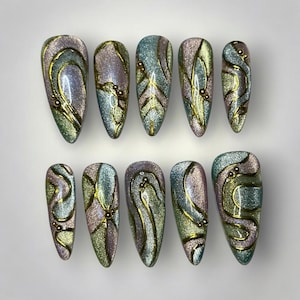 May include: A set of ten almond-shaped false nails. Each nail has a shimmering, iridescent finish in green, purple, and gold. The design includes gold swirls and small gold bead accents, creating a glamorous and artistic look.