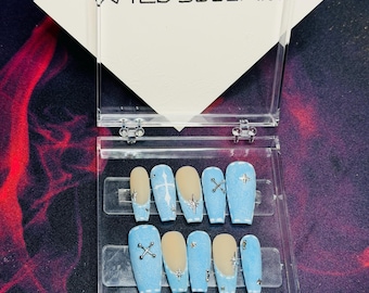 May include: A set of coffin-shaped false nails in a clear plastic case. The nails are light blue with silver accents, including crosses and starbursts. Some nails have a nude-colored base. The case has the text "WILD SOUL ART" on a white background.