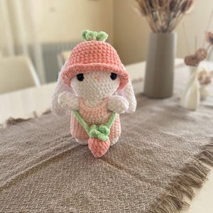 Könnte beinhalten: Handgefertigte Häkelkaninchenpuppe mit rosa Hut mit grünen Blättern und rosa Kleid mit Erdbeer-Tasche. Das Kaninchen hat weißes Fell, schwarze Augen und lange Ohren. Die Puppe sitzt auf einer braunen, strukturierten Oberfläche.