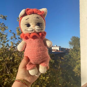 May include: A handmade crochet cat doll in a pink outfit with a matching bonnet. The doll has a cream-colored face, ears, and paws, with black eyes and whiskers. The doll is approximately 8 inches tall.