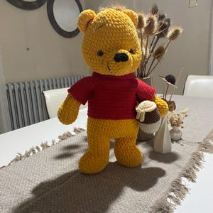 Könnte beinhalten: Ein handgefertigter, gelber Häkelbär mit rotem Hemd, der einen Honigtopf hält. Der Bär hat eine schwarze Nase und steht auf einem Tisch mit einem dekorativen Tischläufer. Der Bär ist etwa 30 cm groß.