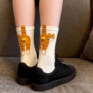 Può includere: Calzini bianchi con un disegno di gatto dei cartoni animati. I calzini presentano un disegno di gatto a strisce arancioni e bianche sul retro, con la coda e il posteriore del gatto visibili. Indossati con scarpe da ginnastica nere.