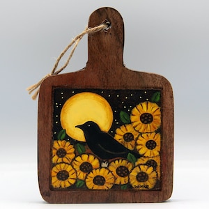 May include: A wooden cutting board ornament with a handle and a jute twine loop. The surface features a hand-painted scene of a black bird perched among yellow sunflowers against a dark background with a large yellow moon and the signature E. Witke.