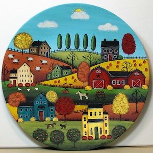 May include: A circular painting depicting a rural landscape with houses, a red barn, sheep, trees in autumn colors, pumpkins, a horse-drawn cart, a dog, and a cat. The scene features a blue sky with a sun and clouds, signed S. Wittke at the bottom.