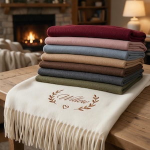 Könnte beinhalten: Ein Stapel gefalteter Fleecedecken in verschiedenen Farben wie Burgunderrot, Rosa, Blau, Beige, Braun, Anthrazit und Oliv, gekrönt von einer cremefarbenen Decke mit dem gestickten Namen Willow, einem Herz und Blattzweigen sowie Fransen.