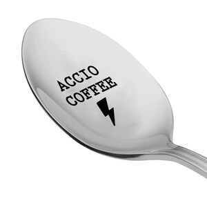 May include: A silver spoon with the words "ACCIO COFFEE" and a black lightning bolt graphic. The spoon is angled, showing the curved bowl and the handle.