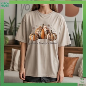 May include: Beige t-shirt featuring a pumpkin graphic with the words "grateful thankful blessed". The pumpkins have orange, brown, and leopard print patterns. The shirt has short sleeves and a crew neck.