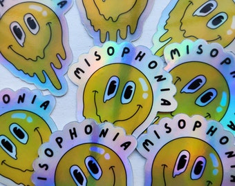 May include: A collection of holographic stickers featuring a yellow, smiling face with the word "MISOPHONIA" in a circular border. The design has a dripping effect, and the stickers have a shiny, iridescent finish.